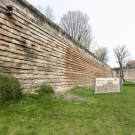 Das Bild zeigt eine große Steinmauer mit horizontal geschichteten Steinen. Davor befindet sich eine Grasfläche mit einigen Büschen und kleinen gelben Blumen sowie eine Informationstafel, die vermutlich historische oder erklärende Inhalte zeigt. Im Hintergrund sind kahle Bäume und ein Weg zu sehen, der in einen anderen Bereich führt, unter einem bewölkten Himmel.