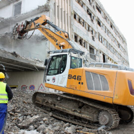 Das Bild zeigt eine Baustelle, auf der ein großer Bagger ein Gebäude abreißt. Der Bagger ist mit einem hydraulischen Abbruchhammer ausgestattet, der Betonteile des Gebäudes zerkleinert. Ein Bauarbeiter mit gelbem Helm und Warnweste steht in der Nähe und beobachtet den Abbruch.