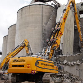 Auf dem Bild ist eine Baustelle zu sehen, auf der zwei große Bagger ein massives Betonbauwerk abreißen. Im Vordergrund steht ein Arbeiter in grüner Jacke nahe eines der Bagger. Im Hintergrund sind beschädigte zylindrische Betonstrukturen zu erkennen, vermutlich Silos.