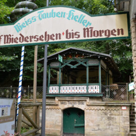 Auf dem Bild ist ein Braukeller zu sehen. Die Umgebung ist von Bäumen umgeben, mit Holzgeländern und einer kleinen Struktur mit grünem Eingang, die auf den Braukeller hinweist.