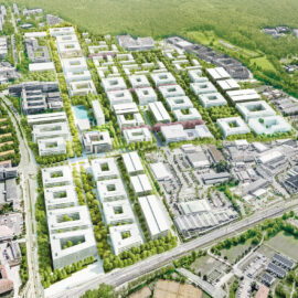 Das Bild zeigt eine Luftaufnahme eines großen städtischen Areals mit zahlreichen modernen, überwiegend weißen Gebäuden, die in einem rasterartigen Muster angeordnet sind. Zwischen den Gebäuden befinden sich Grünflächen mit Bäumen und Rasen, sowie Straßen, Wege und Parkplätze. Im Hintergrund sind weitere Bebauung und ein bewaldetes Gebiet zu erkennen.