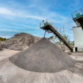 Das Bild zeigt ein Industriegelände mit einem großen Kies- oder Steinhaufen in der Mitte. Zwei erhöhte Förderbänder, gestützt von Metallkonstruktionen, verlaufen links und rechts und dienen dem Materialtransport. Im Hintergrund ist ein großer Erd- oder Felshügel zu sehen unter einem teils bewölkten Himmel.