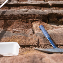 Das Bild zeigt eine Nahaufnahme einer Gesteinsoberfläche mit deutlich erkennbaren Schichten. Links ist ein Maßband senkrecht angebracht, um die Größenverhältnisse der Gesteinsschichten zu verdeutlichen. Rechts liegt ein Geologiehammer mit blauem Griff auf einer der Schichten, unten links befindet sich ein weißer Kunststoffbehälter.