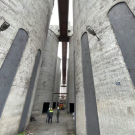 Auf dem Bild ist eine schmale Gasse zwischen großen, zylindrischen Betonbauten zu sehen. Zwei Personen stehen im Vordergrund, weiter hinten ist ein Fahrzeug erkennbar. Über den Bauwerken verlaufen Metallstege, und am Boden sind Pflastersteine sowie etwas Grün zu sehen.