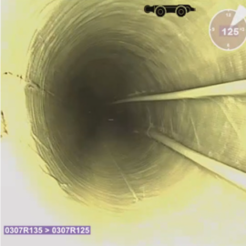 Das Bild zeigt die Innenansicht eines runden Rohres, aufgenommen mit einer Inspektionskamera. Die Rohrwand ist gleichmäßig strukturiert und verläuft in die Tiefe, wodurch ein Tunnel-Effekt entsteht. Auf der rechten Seite sind zwei parallel verlaufende Leitungen oder Kabel sichtbar, und oben links befindet sich eine grafische Anzeige. Unten links ist ein kleiner Textbereich mit Zahlen und Buchstaben eingeblendet, der vermutlich die Position oder den Abschnitt des Rohres angibt.