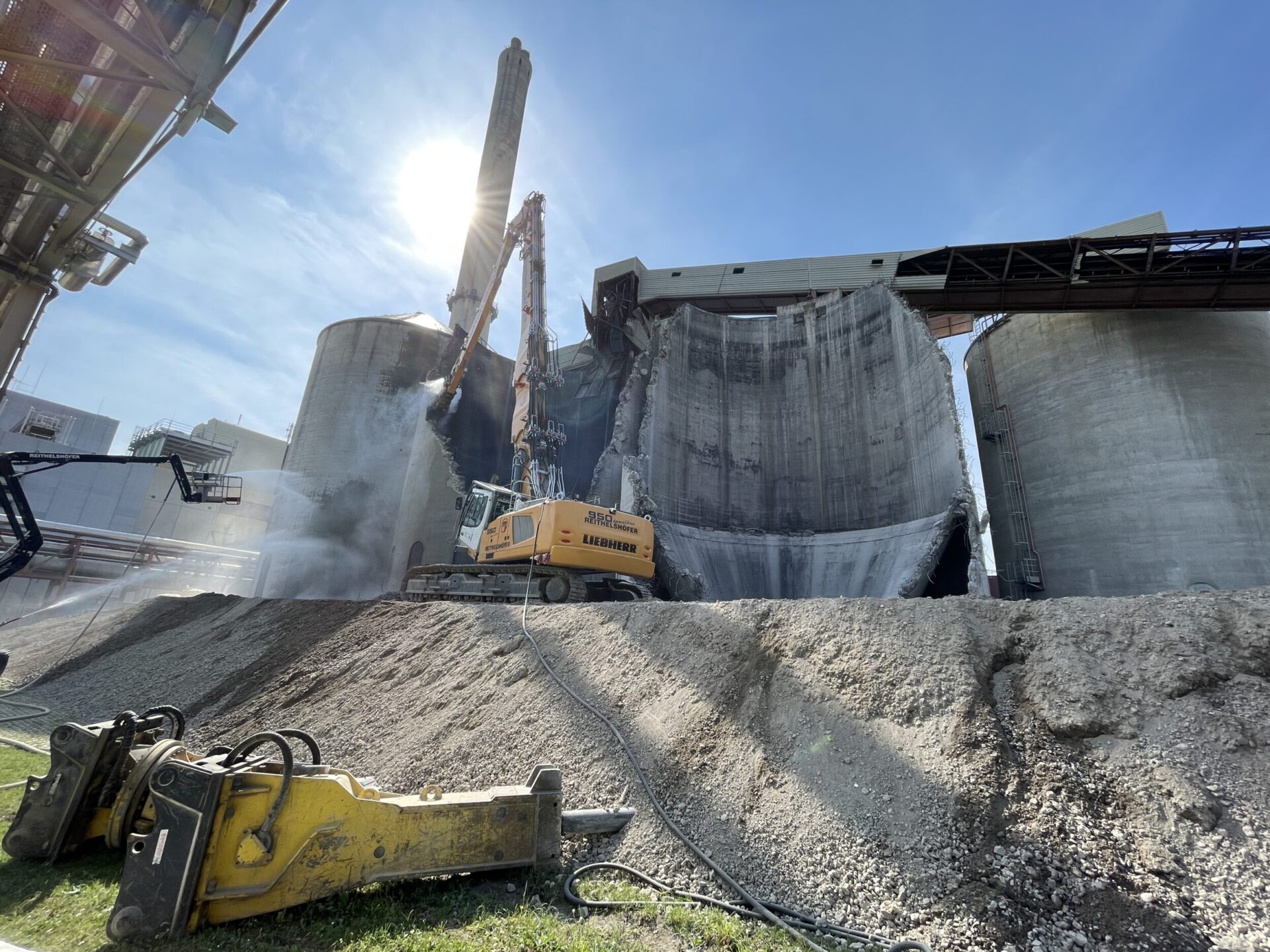 Auf dem Bild ist eine Baustelle mit großen, zylindrischen Betonbauten zu sehen. Ein gelber Bagger mit Hydraulikhammer bearbeitet eines der Bauwerke, das bereits teilweise abgetragen ist. Im Vordergrund liegen weitere Baumaschinenteile auf einem Schotterhaufen, während die Sonne hell über der Szene scheint.