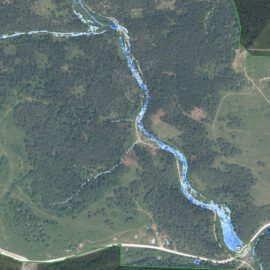 Das Bild zeigt eine Luftaufnahme einer Landschaft mit einem sich schlängelnden Fluss, der in Blau hervorgehoben ist und von der Bildmitte oben nach rechts unten verläuft. Die Umgebung besteht aus dichten Wäldern, offenen Feldern und vereinzelt stehenden Gebäuden. Straßen sind ebenfalls sichtbar, darunter eine markante Straße im unteren Bereich des Bildes und eine weitere, die diese im rechten Bereich kreuzt.