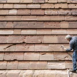 Eine Person arbeitet an einer großen Wand aus rechteckigen Steinplatten in verschiedenen Braun- und Beigetönen. Die Person hält einen Hammer und scheint die Platten zu überprüfen oder zu reparieren. Einige der Steine zeigen sichtbare Abnutzungs- oder Schadensspuren.