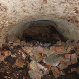 Das Bild zeigt einen unterirdischen Tunnel oder Keller mit einer gewölbten Decke aus Ziegelsteinen. Auf dem Boden liegt ein großer Haufen aus Schutt, bestehend aus zerbrochenen Ziegeln, Steinen und weiteren Trümmern.