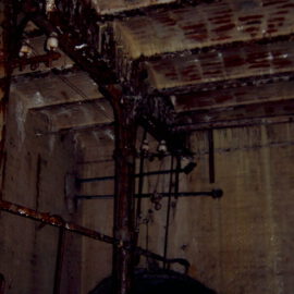 Das Bild zeigt einen alten, verrosteten Keller- oder Maschinenraum mit Metallrohren und Trägern. Wände und Decke bestehen aus Beton und weisen deutliche Wasserschäden und Rostflecken auf. Verschiedene Rohre, Ketten und mechanische Bauteile hängen von der Decke oder sind an den Wänden befestigt.