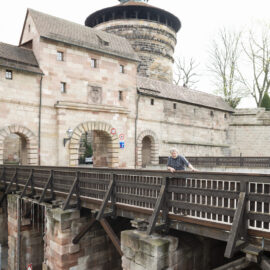 Das Bild zeigt ein historisches Steingebäude mit einem Turm, vermutlich Teil einer Burg oder Befestigungsanlage. Eine Holzbrücke führt zu gewölbten Eingängen, an denen Verkehrsschilder für Fußgänger und Fahrzeuge angebracht sind. Eine Person lehnt am Brückengeländer und blickt nach unten, im Hintergrund stehen laublose Bäume.