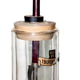 Das Bild zeigt ein Laborgerät mit einem gläsernen Behälter, der auf einem Holzsockel steht und oben ebenfalls mit Holz eingefasst ist. Im Inneren befindet sich eine Metallstange, die mit einem verstellbaren Armmechanismus am oberen Teil verbunden ist.