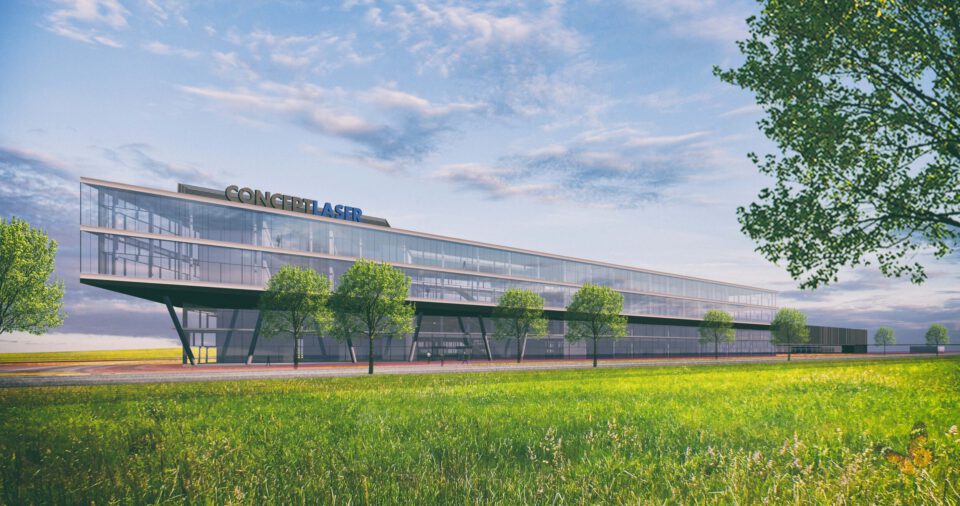 Das Bild zeigt ein modernes, mehrstöckiges Gebäude mit großen Glasfenstern und einer klaren, geradlinigen Architektur. Vor dem Gebäude erstreckt sich eine gepflegte Rasenfläche mit mehreren Bäumen. Der Himmel ist teils bewölkt mit sichtbaren blauen Abschnitten.