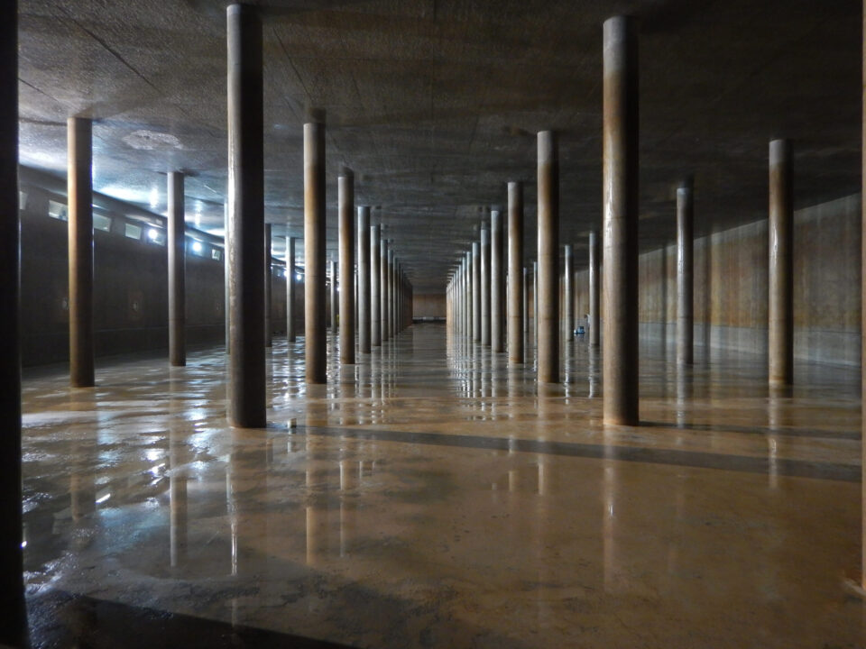 Das Bild zeigt eine unterirdische Struktur mit vielen senkrechten Säulen, die eine flache Decke tragen. Der Boden ist nass und spiegelt die Säulen sowie die Decke wider.