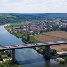 Das Bild zeigt eine Luftaufnahme eines Flusses, der sich durch eine Landschaft mit grünen Hügeln und Feldern schlängelt. Eine Brücke überquert den Fluss, auf der mehrere Fahrzeuge unterwegs sind. Auf einer Seite des Flusses liegt eine Stadt mit vielen Häusern und Gebäuden, umgeben von landwirtschaftlichen Flächen und Waldstücken.