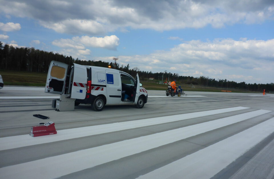 Das Bild zeigt eine Szene auf einer großen asphaltierten Fläche, vermutlich einer Start- oder Landebahn. Ein weißer Transporter mit geöffnetem Heck steht am Rand, während eine Person in orangefarbener Jacke weiter entfernt an einer Maschine arbeitet. Im Hintergrund sind Bäume und ein teils bewölkter Himmel zu sehen.