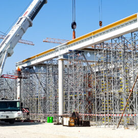Das Bild zeigt eine Baustelle, auf der ein großer Kran einen Stahlträger anhebt. Im Hintergrund sind mehrere Gerüstkonstruktionen und weitere Kräne zu sehen. Der Boden ist mit Kies bedeckt, und verschiedene Baumaterialien sowie Geräte liegen verteilt auf dem Gelände.