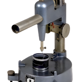 Das Bild zeigt ein Präzisionsmessgerät, vermutlich ein Höhenmessgerät oder eine Messuhr. Es steht auf einem zylindrischen Sockel mit drei verstellbaren Füßen zur Nivellierung. Eine senkrechte Säule mit Skala und ein verstellbarer Arm mit Messfühler ermöglichen genaue Höhen- oder Maßbestimmungen eines darunterliegenden Objekts.