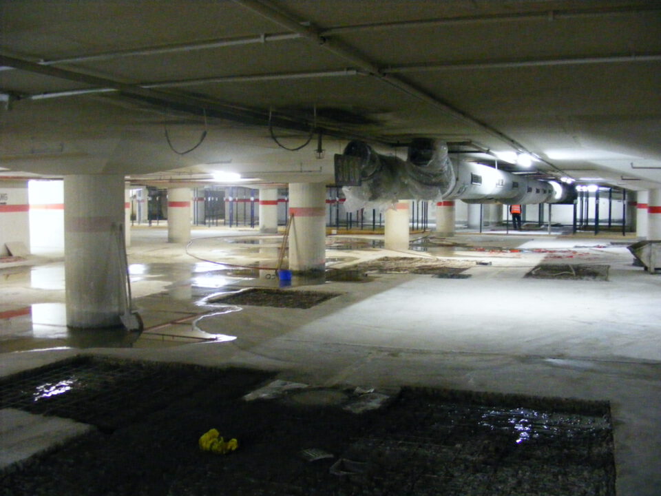 Das Bild zeigt eine Tiefgarage oder einen Kellerbereich, der sich im Bau oder in der Sanierung befindet. Mehrere Betonsäulen stützen die Decke, an der große Lüftungskanäle verlaufen. Auf dem teilweise nassen Boden sind Pfützen, freigelegter Untergrund sowie Baugeräte und Materialien verteilt.