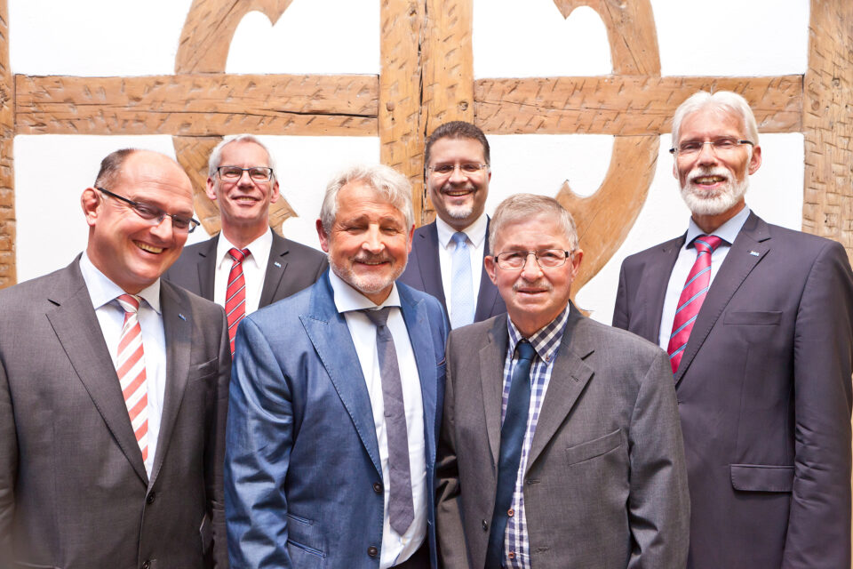 Sechs Männer in formeller Kleidung stehen gemeinsam für ein Foto. Im Hintergrund sind Holzbalken und eine weiße Wand zu sehen.