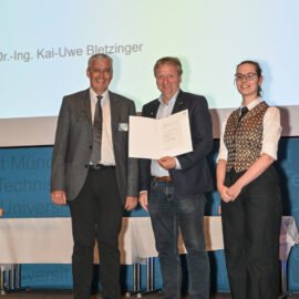 Drei Personen stehen auf einer Bühne, die mittlere Person hält ein Dokument oder eine Urkunde in den Händen. Im Hintergrund ist eine Projektion mit Text zu sehen, die auf eine Präsentation oder Auszeichnung hinweist.