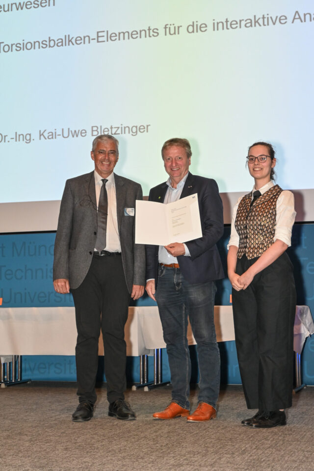 Drei Personen stehen auf einer Bühne, die mittlere Person hält ein Dokument oder eine Urkunde in den Händen. Im Hintergrund ist eine Projektion mit Text zu sehen, die auf eine Präsentation oder Auszeichnung hinweist.