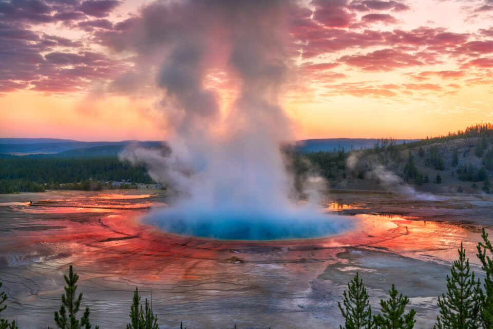 Das Bild zeigt eine geothermische heiße Quelle, aus deren Mitte Dampf aufsteigt. Das Wasser ist in der Mitte leuchtend blau und von rot-orangenen Farbtönen am Rand umgeben. Im Hintergrund sind Bäume, Hügel und ein farbenfroher Himmel bei Sonnenauf- oder -untergang zu sehen.