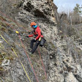 Das Bild zeigt eine Person in Kletterausrüstung, die an einer steilen Felswand arbeitet. Die Person trägt einen orangefarbenen Helm, eine rote Jacke und ist mit mehreren farbigen Seilen gesichert, die an einem Netz befestigt sind, das die Felswand stabilisiert. Im Hintergrund sind felsige Strukturen und ein Drahtzaun zu sehen, was auf eine Sicherungsmaßnahme in einem Hangbereich hinweist.
