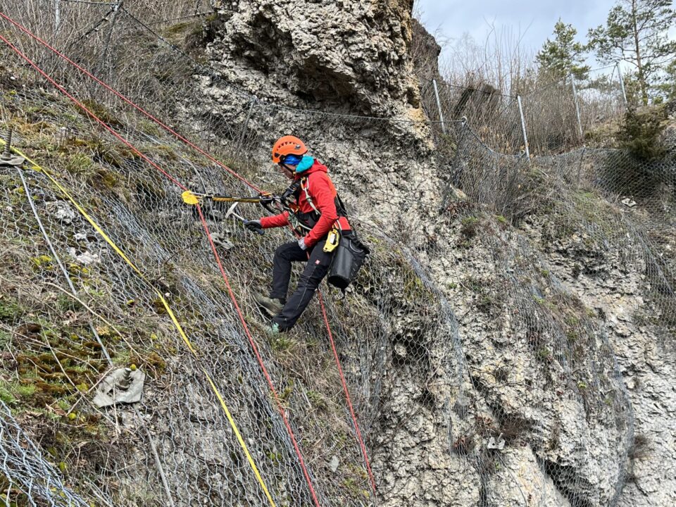 Das Bild zeigt eine Person in Kletterausrüstung, die an einer steilen Felswand arbeitet. Die Person trägt einen orangefarbenen Helm, eine rote Jacke und ist mit mehreren farbigen Seilen gesichert, die an einem Netz befestigt sind, das die Felswand stabilisiert. Im Hintergrund sind felsige Strukturen und ein Drahtzaun zu sehen, was auf eine Sicherungsmaßnahme in einem Hangbereich hinweist.