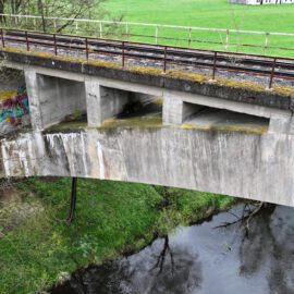 Das Bild zeigt eine zweistöckige Betonbrücke, wobei die obere Ebene mit Bahngleisen und einem Metallgeländer ausgestattet ist. Die untere Ebene ist eine Bogenbrücke, die über einen Fluss führt. Umgeben ist die Brücke von grüner Wiese und Bäumen.