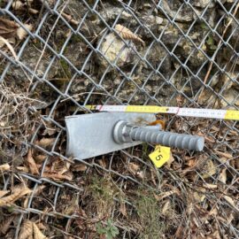 Das Bild zeigt ein Detail einer Sicherungskonstruktion an einer Felswand. Im Vordergrund ist ein massiver Metallanker mit geriffeltem Bolzen zu sehen, der durch ein Drahtnetz in den Fels eingebaut ist. Daneben liegt ein gelbes Markierungsschild mit einer Zahl sowie ein Maßband, das zur Vermessung der Position dient. Die Umgebung besteht aus Steinen, trockenen Blättern und etwas Moos.