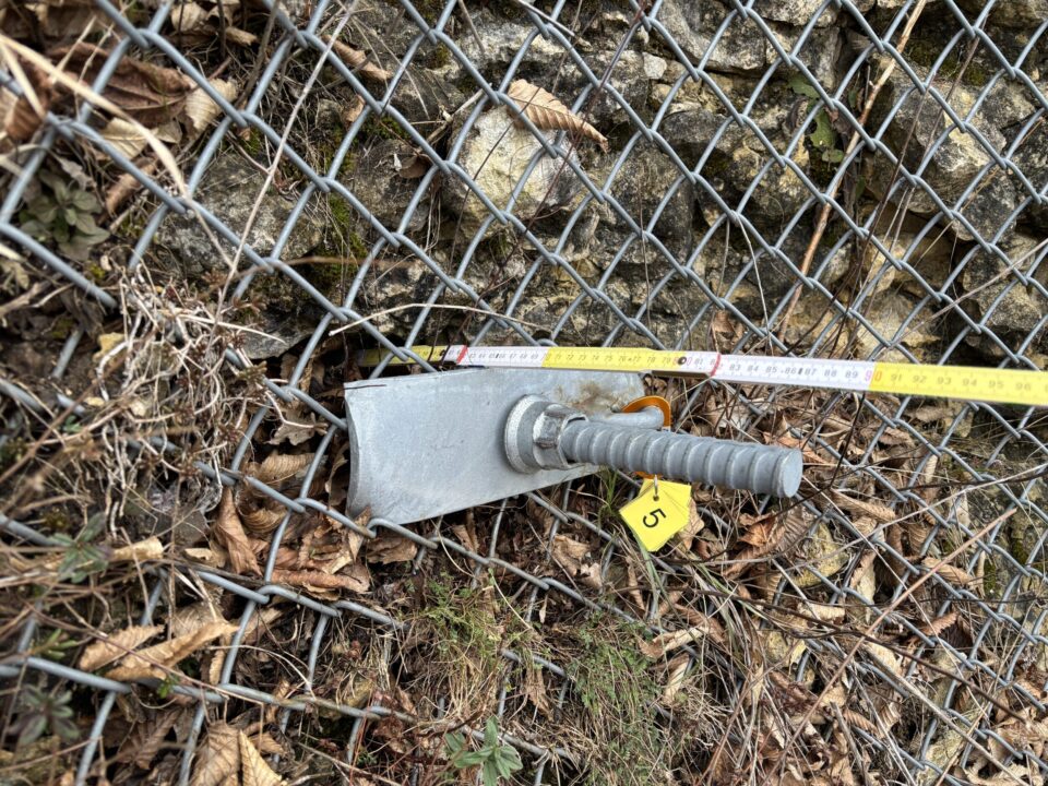 Das Bild zeigt ein Detail einer Sicherungskonstruktion an einer Felswand. Im Vordergrund ist ein massiver Metallanker mit geriffeltem Bolzen zu sehen, der durch ein Drahtnetz in den Fels eingebaut ist. Daneben liegt ein gelbes Markierungsschild mit einer Zahl sowie ein Maßband, das zur Vermessung der Position dient. Die Umgebung besteht aus Steinen, trockenen Blättern und etwas Moos.