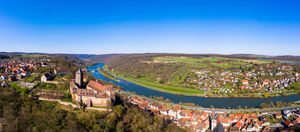 Das Bild zeigt eine Luftaufnahme einer malerischen Landschaft mit einem Fluss, der sich durch ein Tal schlängelt. Links vom Fluss liegt eine historische Burg mit roten Ziegeldächern und Steinmauern, umgeben von Bäumen und Grünflächen. Daneben befindet sich eine kleine Stadt mit zahlreichen Häusern, ebenfalls mit roten Dächern.