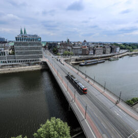 Das Bild zeigt eine Luftaufnahme einer Stadt mit einem Fluss, der sich durch das Stadtgebiet zieht. Eine Brücke überquert den Fluss, auf der eine Straßenbahn oder ein Zug unterwegs ist. Die Stadt vereint moderne Gebäude mit grünen Türmen und ältere Architektur, der Himmel ist teilweise bewölkt.