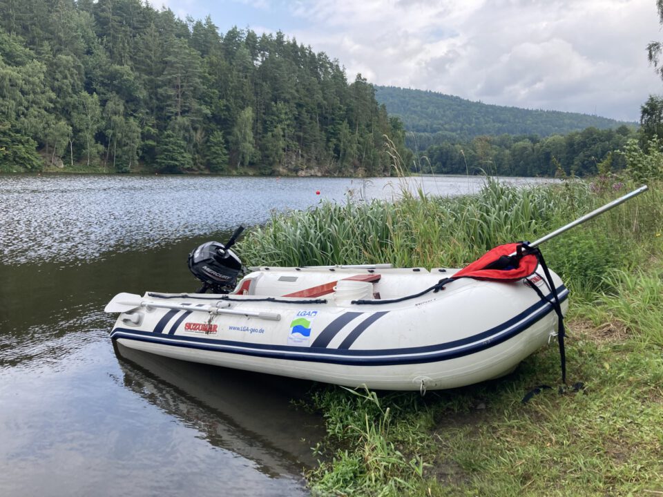 Ein aufblasbares Boot mit Motor liegt teils im Wasser, teils am Ufer eines Sees oder Flusses. Im Hintergrund sind bewaldete Uferbereiche und sanfte Hügel zu sehen.