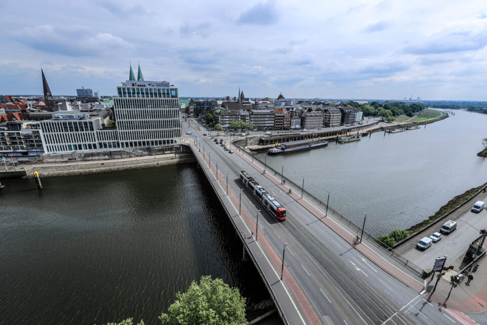 Das Bild zeigt eine Luftaufnahme einer Stadt mit einem Fluss, der sich durch das Stadtgebiet zieht. Eine Brücke überquert den Fluss, auf der eine Straßenbahn oder ein Zug unterwegs ist. Die Stadt vereint moderne Gebäude mit grünen Türmen und ältere Architektur, der Himmel ist teilweise bewölkt.