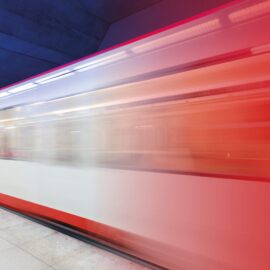 Das Bild zeigt eine U-Bahn in Bewegung, aufgenommen in einer Station. Der Zug ist stark verwischt, was die Geschwindigkeit und Dynamik der Aufnahme betont, und die Farben verlaufen von Weiß im unteren Bereich zu kräftigem Rot im oberen Bereich. Der Bahnsteig ist hell gefliest, während die Wand und Decke der Station in einem satten Blauton gehalten sind, was einen starken Farbkontrast zum Zug erzeugt. Die Beleuchtung ist gleichmäßig und unterstreicht die moderne Atmosphäre der Szene.