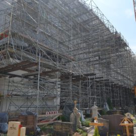 Das Bild zeigt eine große Baustelle mit umfangreichem Gerüst, das die gesamte Seite eines Gebäudes bedeckt. Rechts im Bild steht ein Baukran, und im Vordergrund befinden sich mehrere Grabsteine und Gräber, was auf eine Lage innerhalb oder neben einem Friedhof hinweist.