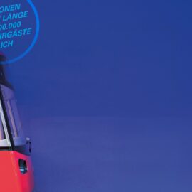 Das Bild zeigt die Front eines roten U-Bahn-Wagens, der vor einem einfarbig blauen Hintergrund positioniert ist. Der Zug ist nur teilweise sichtbar, wobei die Frontscheibe und ein Teil der Seitenwand erkennbar sind. Im oberen linken Bereich befindet sich ein kreisförmiger grafischer Hinweis in hellblauer Schrift mit Informationen zu 49 Stationen, 38 Kilometern Streckenlänge und über 400.000 Fahrgästen täglich. Die Gestaltung wirkt modern und kontrastreich durch die Kombination von kräftigem Blau und Rot.