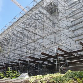 Das Bild zeigt eine große Baustelle mit umfangreichem Metallgerüst, das die sichtbare Seite eines Gebäudes vollständig umgibt und mit weißer Folie abgedeckt ist. Im Hintergrund sind Baukräne zu sehen, die auf laufende Arbeiten hinweisen. Im Vordergrund befinden sich Pflanzen und ein kleines Gebäude mit schrägem Dach.