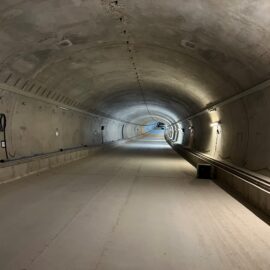 Das Bild zeigt einen neu gebauten Tunnel mit glatten Betonwänden und einer gleichmäßig gewölbten Decke. Der Tunnel ist gut ausgeleuchtet, wobei die Beleuchtung entlang der rechten Wand angebracht ist. Auf beiden Seiten verlaufen Kabel und Leitungen, die teilweise an der Wand befestigt sind, und der Boden ist eben und sauber, ohne Schienen oder Fahrzeuge. Am Ende des Tunnels ist eine leichte Krümmung sichtbar, die in die Ferne führt.