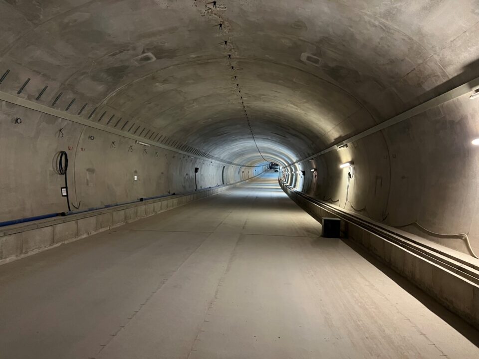 Das Bild zeigt einen neu gebauten Tunnel mit glatten Betonwänden und einer gleichmäßig gewölbten Decke. Der Tunnel ist gut ausgeleuchtet, wobei die Beleuchtung entlang der rechten Wand angebracht ist. Auf beiden Seiten verlaufen Kabel und Leitungen, die teilweise an der Wand befestigt sind, und der Boden ist eben und sauber, ohne Schienen oder Fahrzeuge. Am Ende des Tunnels ist eine leichte Krümmung sichtbar, die in die Ferne führt.