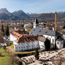 Das Bild zeigt eine Panoramaaufnahme eines Ortes inmitten einer Berglandschaft mit mehreren Gebäuden, darunter eine große Kirche mit hohem Turm und ein angrenzender Friedhof. Ein Baukran steht in der Nähe der Kirche, und im Hintergrund sind bewaldete Hügel sowie schneebedeckte Berge unter blauem Himmel zu sehen. Die Umgebung ist grün und gepflegt mit Wegen und Rasenflächen.