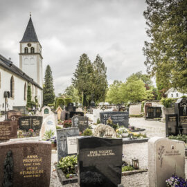 Das Bild zeigt eine Kirche mit hohem Glockenturm auf der linken Seite, umgeben von einem Friedhof. Die Kirche hat weiße Wände und Rundbogenfenster, und im Friedhof stehen zahlreiche Grabsteine und Denkmäler. Bäume und Grünflächen umrahmen die Szene, der Himmel ist wolkenverhangen.