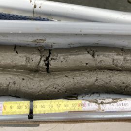 Das Bild zeigt eine Nahaufnahme eines Sedimentkerns aus grauem Schlamm oder Ton in einem zylindrischen Behälter. Unter dem Behälter liegt ein Maßband, das eine Länge des Kerns von etwa 45 bis 70 Zentimeter anzeigt.