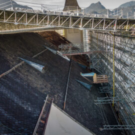 Das Bild zeigt eine Baustelle auf dem Dach eines großen Gebäudes, das mit dunklen Schindeln gedeckt ist. Um das Dach herum sind mehrere Gerüstkonstruktionen aufgebaut, und eine provisorische Brücke aus Metallträgern verläuft quer über das Dach. Im Hintergrund sind Berge unter klarem Himmel zu sehen.
