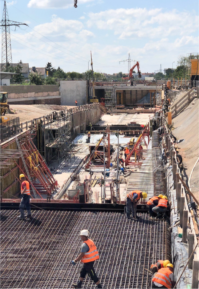 Das Bild zeigt eine große Baustelle für den U-Bahn-Bau, aufgenommen bei Tageslicht. Im Vordergrund arbeiten mehrere Personen in orangefarbenen Sicherheitswesten auf einer Fläche mit dicht verlegtem Bewehrungsstahl. Dahinter sind rote Stahlträger und Schalungselemente zu sehen, die den Tunnelbereich stützen. Im Hintergrund befinden sich weitere Baugeräte, darunter ein Bagger und ein Kran, sowie Betonwände, die den Bauabschnitt begrenzen. Strommasten und ein blauer Himmel mit vereinzelten Wolken runden die Szene ab.