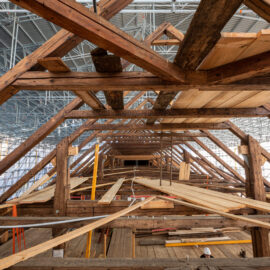 Das Bild zeigt das Innere einer großen Holzkonstruktion, die sich im Bau oder in der Renovierung befindet. Der Blick verläuft durch einen langen Gang aus hölzernen Trägern und Planken, die in einem dreieckigen Muster das Dach stützen. Im Hintergrund sind Gerüste und Baumaterialien wie Holzplanken und Metallstangen zu sehen.