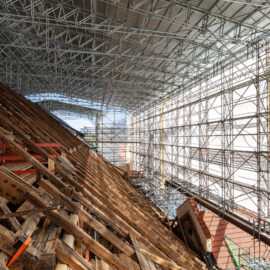 Das Bild zeigt eine große Baustelle im Inneren eines Gebäudes mit umfangreichem Gerüst auf der rechten Seite, bestehend aus Metallträgern in Gitterstruktur. Links befindet sich eine geneigte Holzkonstruktion, vermutlich Teil eines Dachs oder Bodens in Bau oder Sanierung. Eine provisorische Überdachung aus Metall schützt die Baustelle vor Witterungseinflüssen.