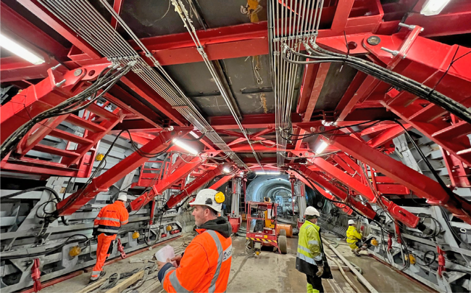 Das Bild zeigt den Innenbereich eines U-Bahn-Tunnels im Bau, der von einer massiven roten Stahlkonstruktion gestützt wird. Die Konstruktion besteht aus zahlreichen Verstrebungen und Trägern, die den Tunnel stabilisieren, während Kabel und Leitungen ordentlich entlang der Decke verlaufen. Mehrere Personen in Sicherheitskleidung und Helmen arbeiten im Tunnel, und im Hintergrund ist ein kleiner Baustellenfahrzeug zu sehen. Die Szene ist gut ausgeleuchtet durch mehrere helle Lampen, die an der Konstruktion befestigt sind.